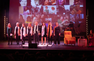 A photograph of performers standing on stage with a collage of photographs projected behind them