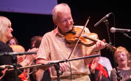 A close up of a man smiling and playing the fiddle as part of a performance
