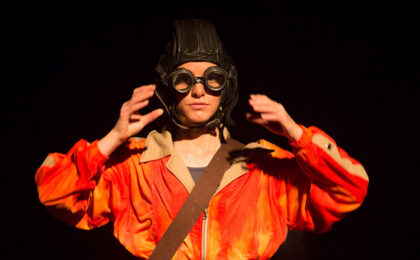 A performer wearing a bright orange bomber jacket and a World War 2 fighter pilot helmet with goggles. 