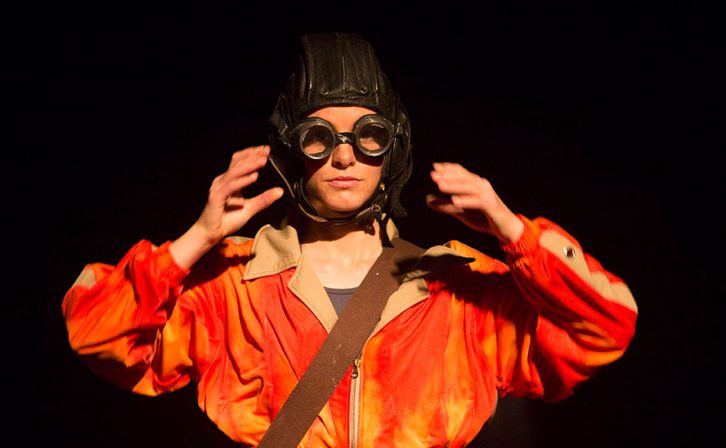 A performer wearing a bright orange bomber jacket and a World War 2 fighter pilot helmet with goggles.  A performer wearing a bright orange bomber jacket and a World War 2 fighter pilot helmet with goggles.