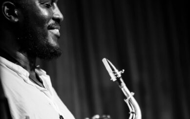 A black and white photo of Tony Kofi smiling and holding his saxophone