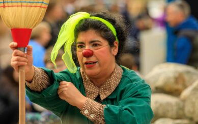 A photo from the performance Juanita's Big Problem, showing a woman holding a broom in front of her. She's wearing a green jackets over a tan shirt and a neon yellow headscarf. On her face is a red clown nose. 