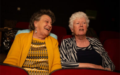 A photograph of two women sitting in theatre seats. The woman on the left is laughing and looking to the woman on the right, who is watching the stage.