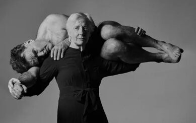 A black and white photograph depicting the dance artist Finola Cronin dressed in black and her arms spread out. Across her arms and shoulders is Mikel Murfi, who is laying on his side and curled around her. 
