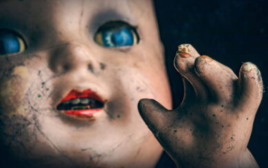 Out of the dark backdrop emerges a face and a hand of a doll with blue eyes and red lips looking into the distance