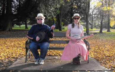 Two people sitting on a park bench playing tennis rackets as guitars