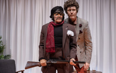A photograph of two actors in stage. In the front is a woman wearing a wig, a fake moustache, glasses and a suit jacket. She's holding a gun. Looking over her shoulder is a man wearing a curly wig and a patterned suit jacket. 