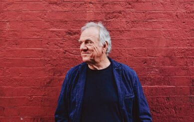 A profile portrait of Ralph McTell wearing a dark T-shirt under a dark denim jacket. He's leaning against a brick wall painted red and smiling. 