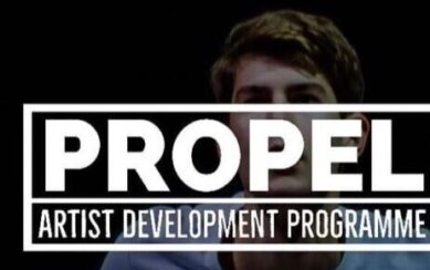The text "Propel Arts Development Program" in white capital letters. The background is a photograph of a man in a light-coloured T-shirt. 