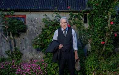 Performer Mikel Murfi wearing a suit andstanding next to a rundown cottage 