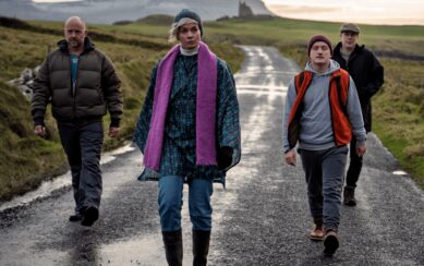 One woman walks ahead while three men follow down an empty road