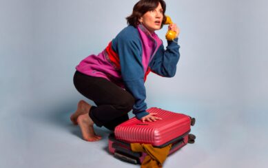 A woman in a colorful fleece jacket crouches over an overstuffed red suitcase while looking startled and holding a vintage yellow telephone receiver