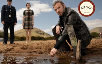 A man in a leather jacket kneels by a shoreline, reaching toward the water while a police officer and a young woman watch from behind.