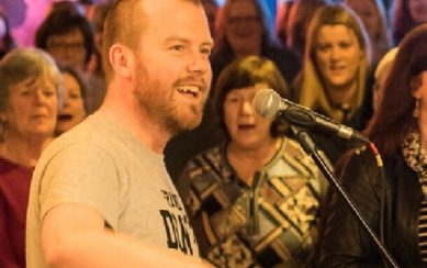 Musician Dave Flynn playing his guitar and singing to an audience with audience in background