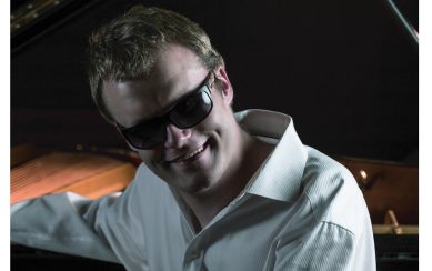 Artist Derek Paravicini sitting at his piano wearing sunglasses 