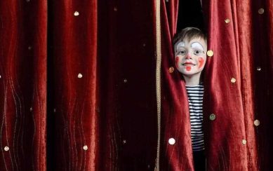 Boy in clown make-up peaking through red curtains 