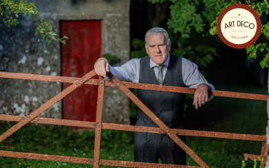 Mikel Murfi in a vest and tie stands by a rusty gate outside a stone building with a red door.