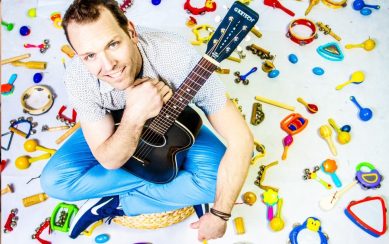 A photo of the artist taking from above with him holding a guitar in his lap while sitting on the ground surrounded by a variety of colourful percussion instruments for kids 