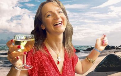 Performer Norma Sheahan wearing a red dress and holding a glass of white wine on a beach with blue sky in the background 