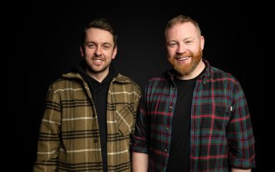 Kendy and Raybo are smiling and wearing plaid shirts in front of a black background