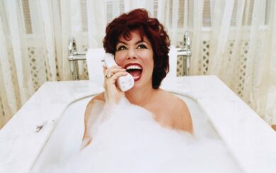 A woman with red hair laughing on a telephone while sitting in a bubble bath.