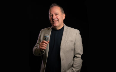 Shane Martin with a microphone on a black background 