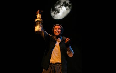 Aidan Dooley stands on stage, holding an oil lamp.