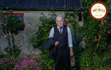 Mikel Murfi, in a vest and tie, stands outside a stone building with flowers around.
