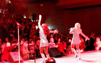 Two singers in sparkly dresses and silver boots perform on stage before a large crowd in red light.