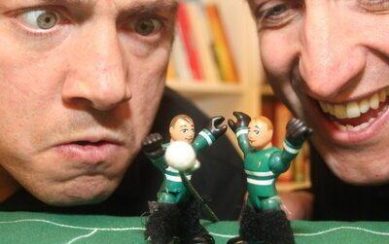 A close-up photograph of two men looking at two Gaelic football player figurines. The man on the left is looking confused, the man on the right is smiling.