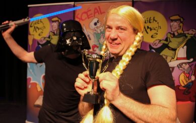 In the foreground is a man wearing a blonde wig with braided pigtails. To his left is a man wearing a Darth Vader mask