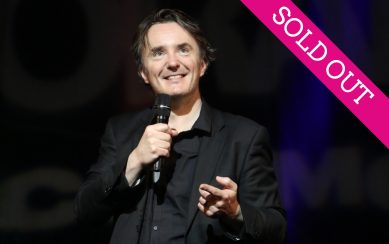 Comedian Dylan Moran standing on stage holding a microphone