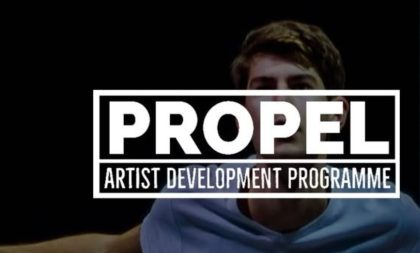 The text "Propel Arts Development Program" in white capital letters. The background is a photograph of a man in a light-coloured T-shirt. The text "Propel Arts Development Program" in white capital letters. The background is a photograph of a man in a light-coloured T-shirt.