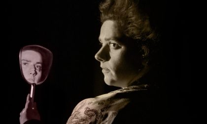 A profile photograph of Sinead Sexton in costume from her play Gazelle. She's holding up a mirror that reflects her face.