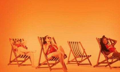 Three girls sunbathing on deck chairs on a blank orange background