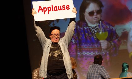 A young man on stage, holding up a sign that says Applause