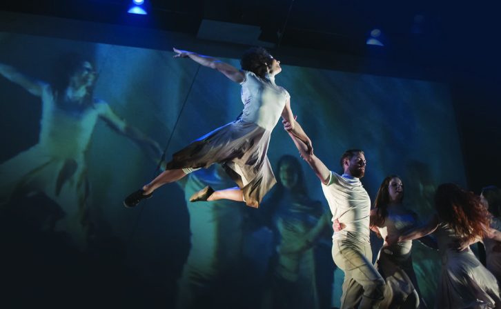 Five dancers on the stage with projected visuals on the back wall and a performer floating above on suspension wires. Five dancers on the stage with projected visuals on the back wall and a performer floating above on suspension wires.