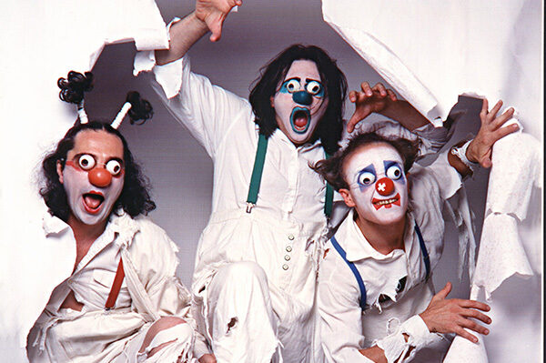 A photograph of three men dressed as clowns, bursting through a white sheet of paper energetically. They're wearing white clothes which have rips, red clown noses and white face paint. Their eyes are covered with white half-spheres with holes in the center.