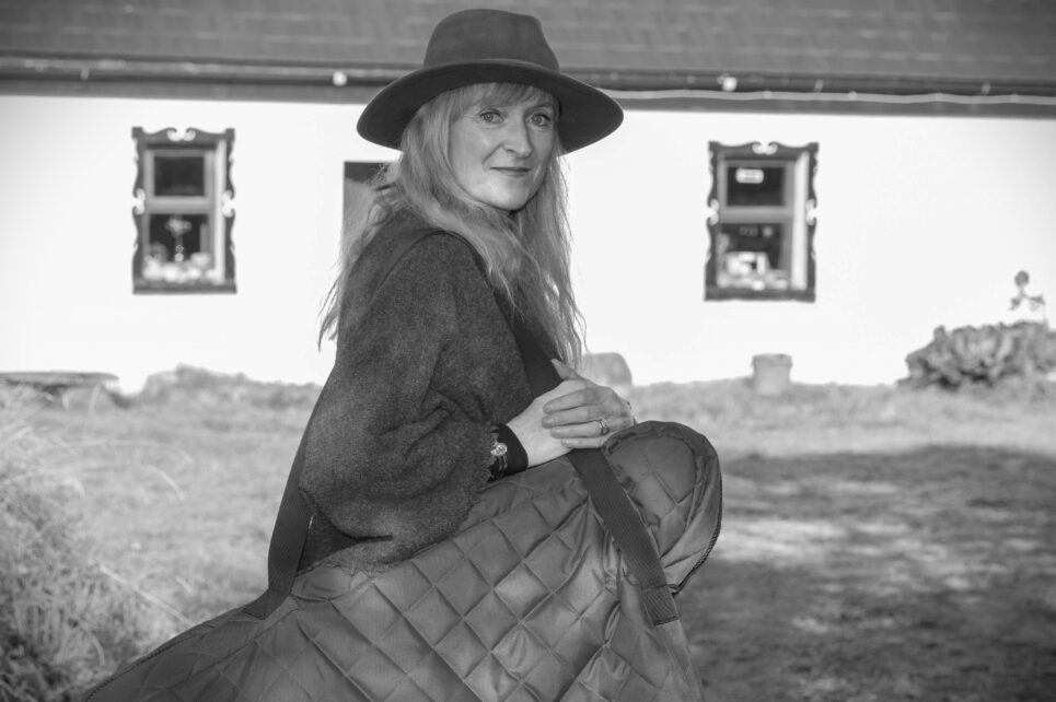 Black and white photograph of artist holding her instrument with cottage in the background