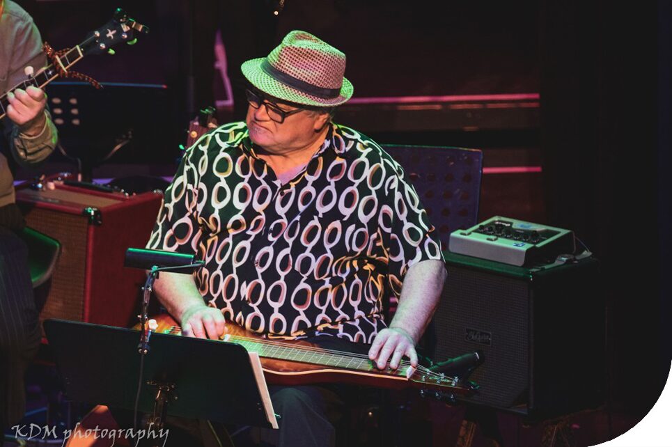 A photograph of Francie Lenehan on stage, wearing a dark shirt with a pattern of white ovals. He's wearing a light coloured fedora hat. He's playing a dulcimer.