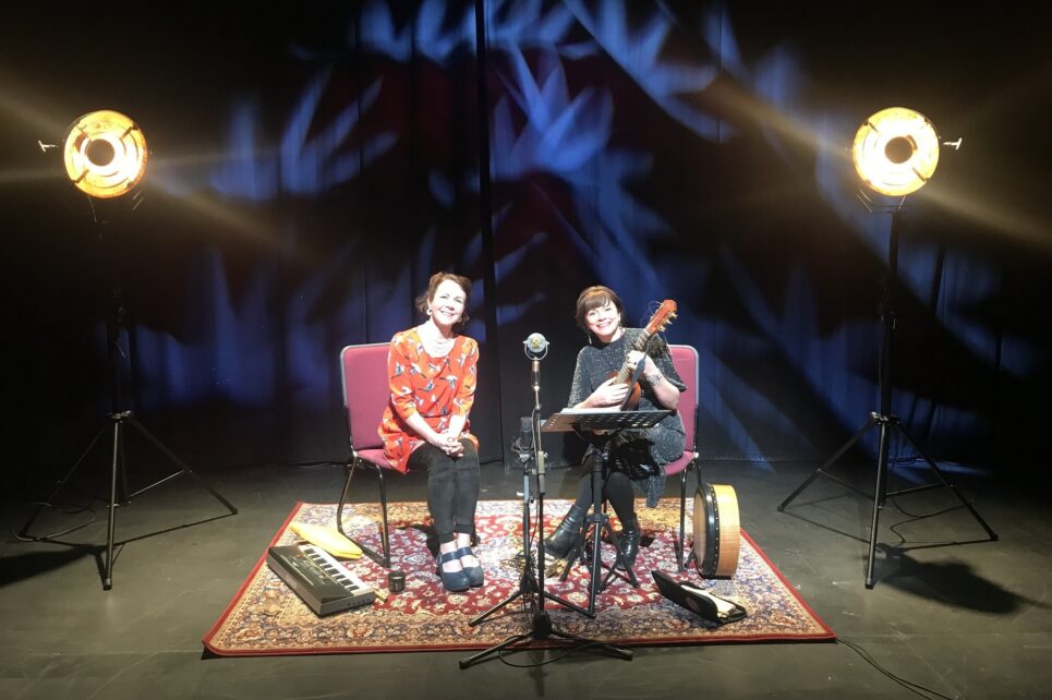 A photograph of Nuala Kennedy and Cathy Jordan sitting on stage under spotlights. Underneath their chairs is a colourful carpet. Nuala is on the left and Cathy on the right, holding a ukulele.