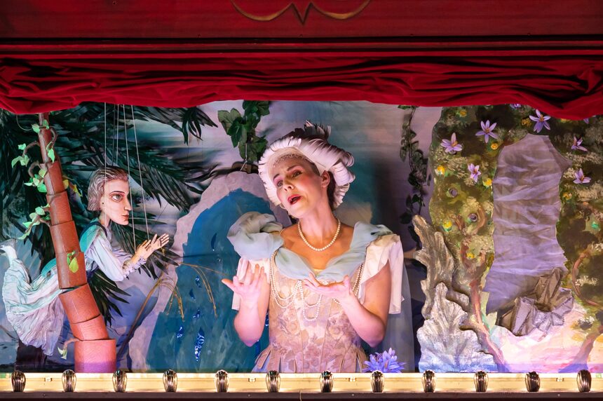 A photograph of an actress in a puppet theatre, singing. Next to her is a wooden puppet resembling her.