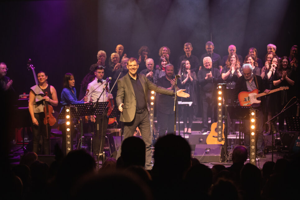 A photograph from the audience's perspective. It depicts Kieran Quinn on stage during a performance. He is dressed in a suit and is holding his arms out toward the audience. Behind him are other musicians and the Theme Night choir. 