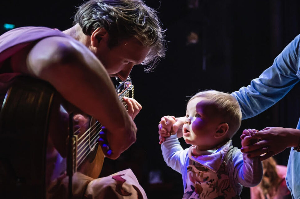 A photograph of a baby listening attentively to a man playing a guitar. 