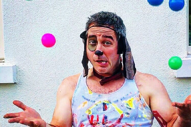 Man wearing a tank top painted over, floppy ears, and his face painted to resemble a dog's. He's juggling multiple balls