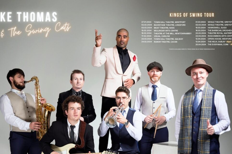A photo of Luke Thomas and The Swing Cats. Luke is standing in the centre in a white suit jacket and black shirt, surrounded by the other musicians