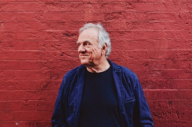 A profile portrait of Ralph McTell wearing a dark T-shirt under a dark denim jacket. He's leaning against a brick wall painted red and smiling. 