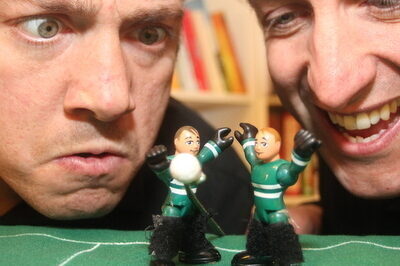A close-up photograph of two men looking at two Gaelic football player figurines. The man on the left is looking confused, the man on the right is smiling. 