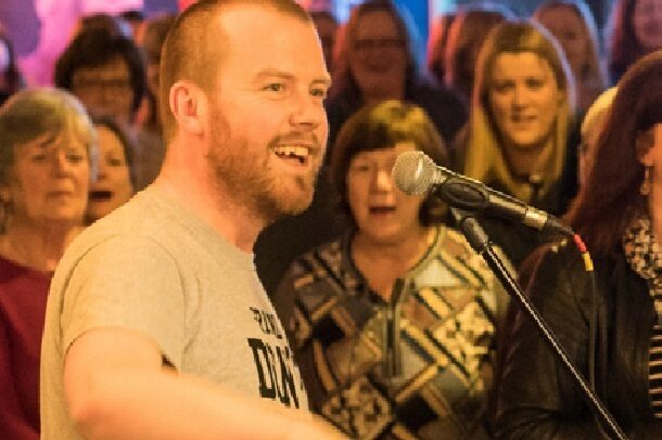 A photograph of Dave Flynn singing in front of a group of people singing along with him. 