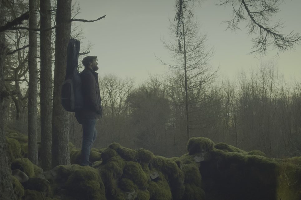 Image of performer standing in a forest with a guitar on his back.